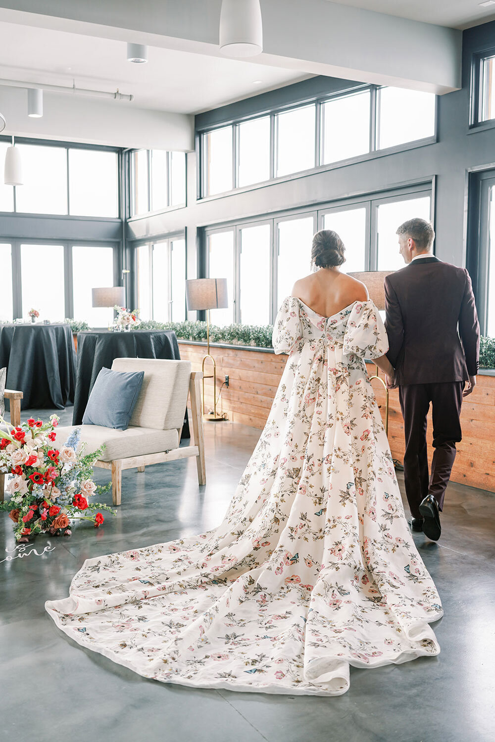 Bride in a floral gown with a long train walking hand in hand with the groom in a burgundy tuxedo during a modern wedding inspiration shoot.