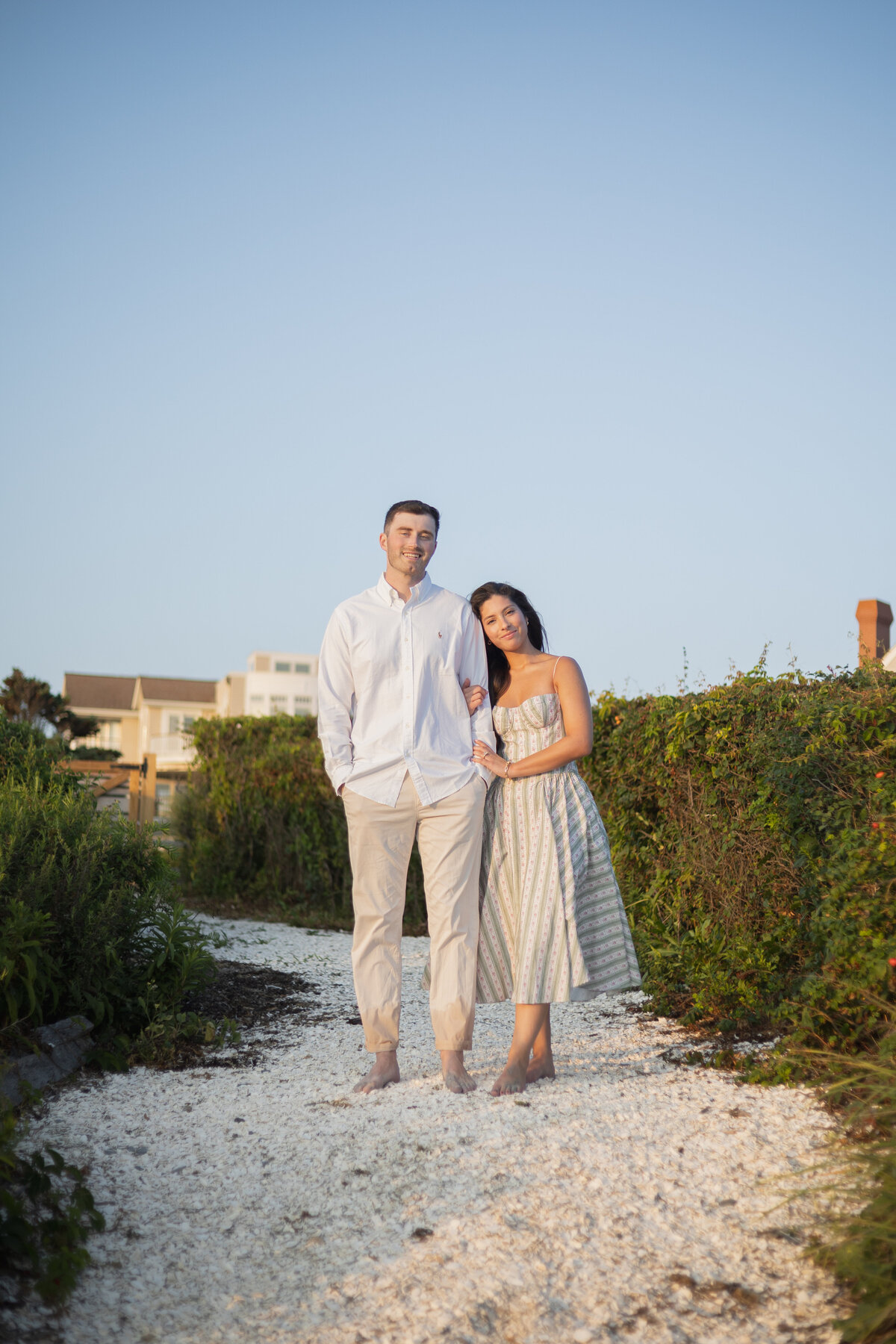 Collins Beach Newport RI | Kelsey Sheehan Photography Timeless Rhode Island Weddings | A couple stands on a pathway surrounded by greenery, with the woman leaning gently on the man's arm. They are barefoot and appear relaxed and happy.