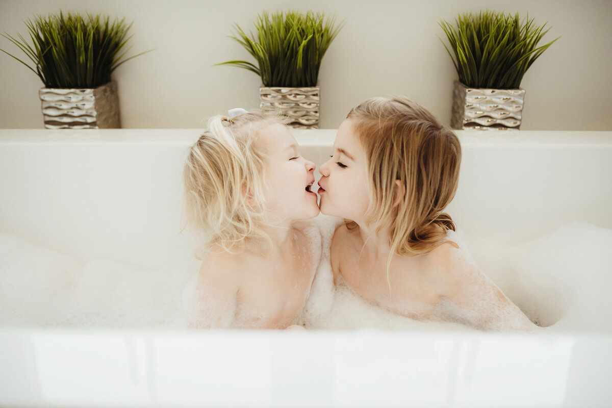 Big sister leaning toward her little sister to kiss her while sitting in the bubble bath.