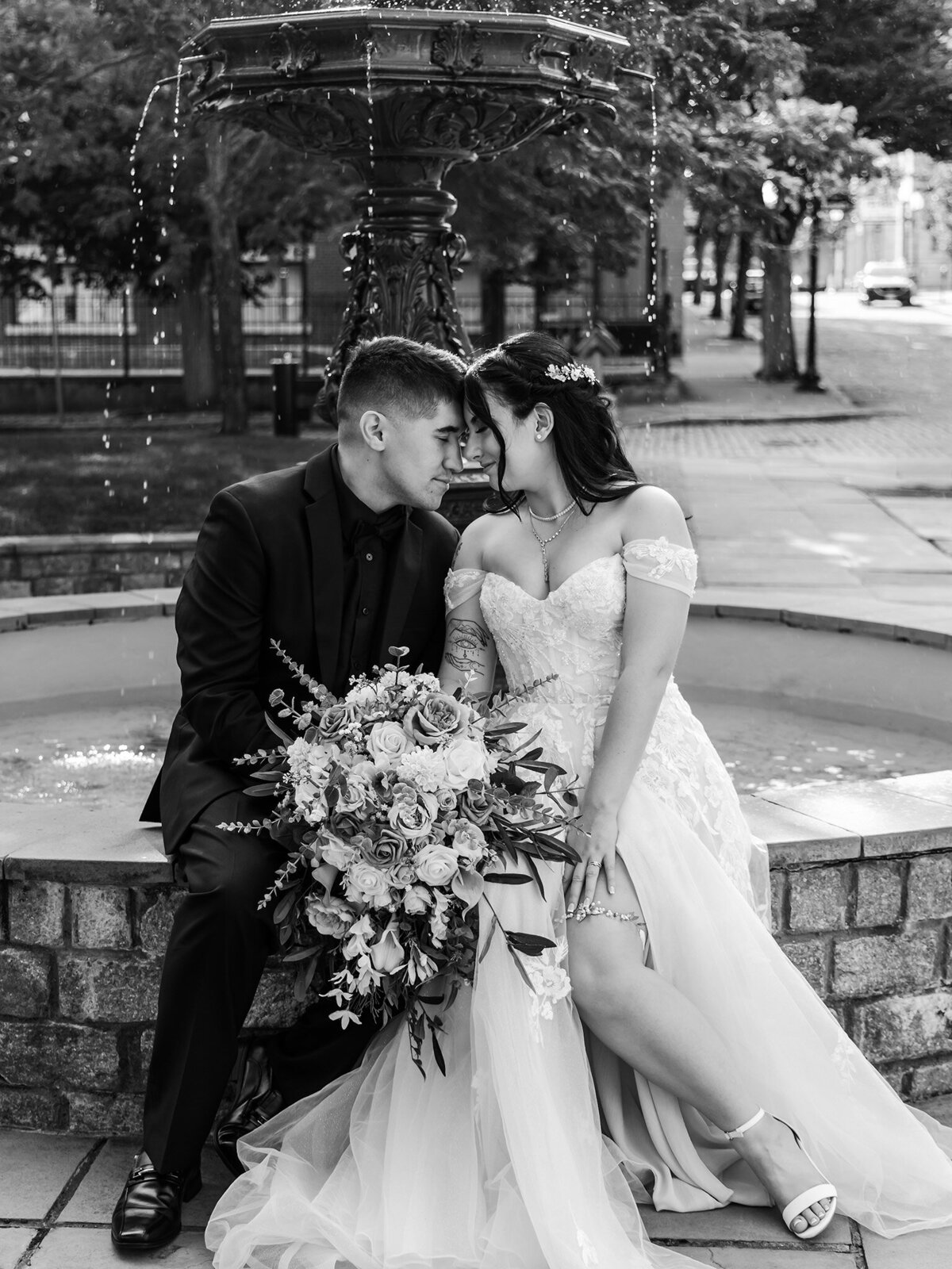 Black and white photograph of a newlywed bride and groom sitting on the edge of a water fountain with the groom wearing an all black tux and the bride wearing an off-the-shoulder white gown with embellished lace corset and a-line skirt holding her cascading wedding bouquet  in the middle of their laps while they put their heads together