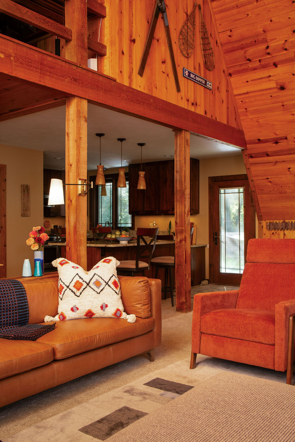 A warmly lit, open-plan wood cabin living room and kitchen area. A brown leather sofa with patterned pillows and an orange armchair sit on a neutral rug. Wooden beams and posts support the structure. Pendant lights hang over a kitchen counter with bar stools. Rustic decor, including snowshoes and tools, adorn the wood-paneled walls.