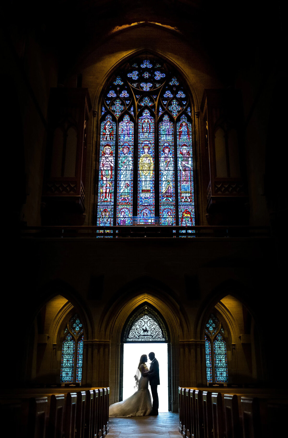 Silhouette of a bride and groom, inside the doorway of Bryn Athyn Cathedral, under a large stained glass window