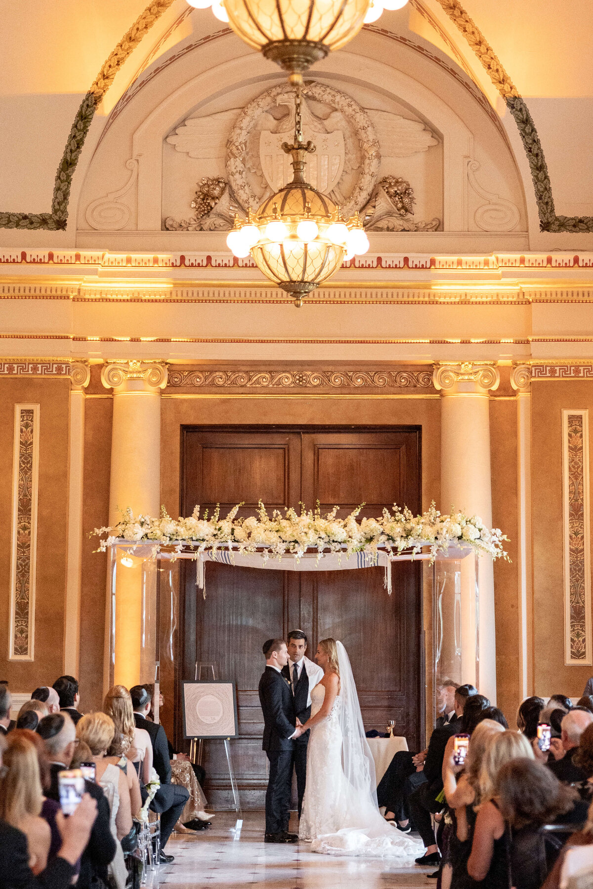 A bride and groom stand together under a floral arch at the altar during a wedding ceremony. Guests are seated on both sides in an elegant hall with high ceilings and ornate decor.