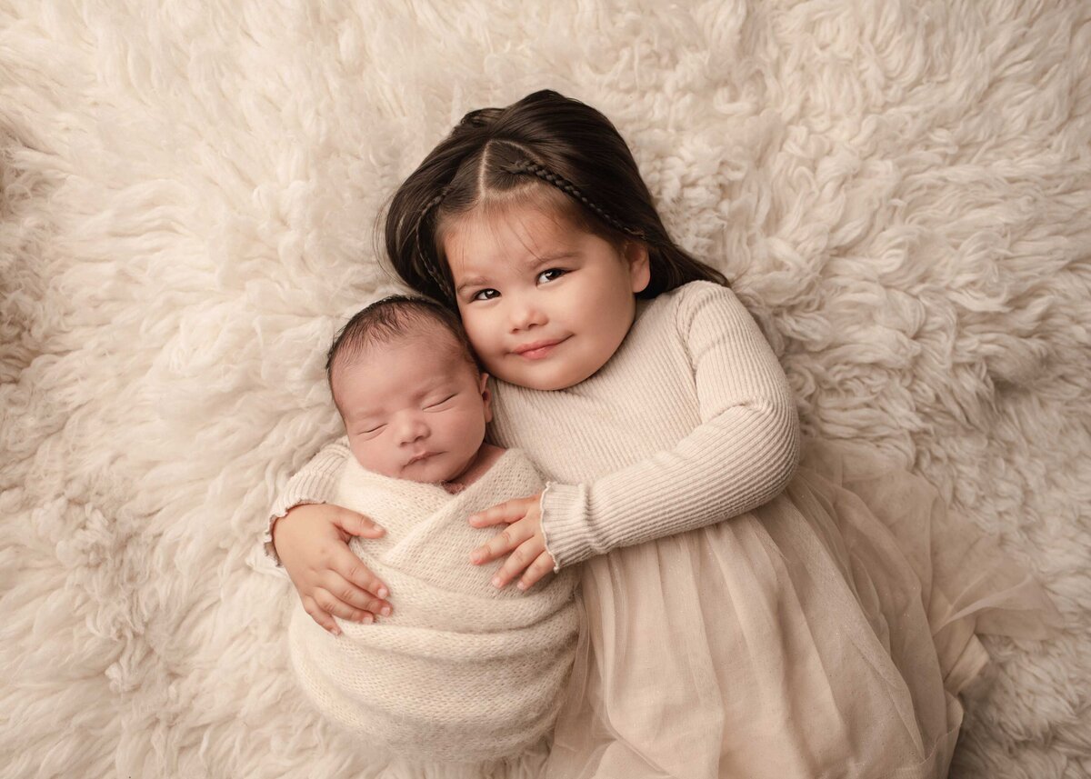 Aerial image of Murrieta newborn photoshoot. Toddler sister has her newborn baby brother resting on her arm. Her cheek is gently resting on her brother's temple. She is smiling at the camera. Captured by best Murrieta newborn photographer Bonny Lynn Photography