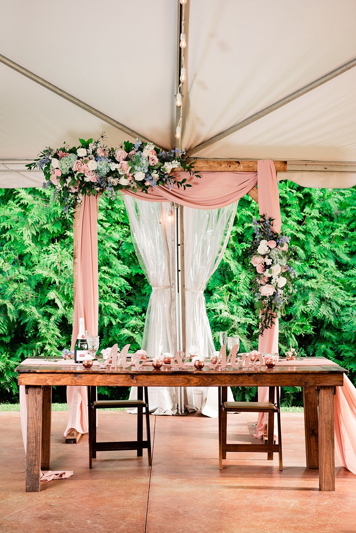 A barn wood farm table sits under a white wedding tent with a wooden arbor behind the table. The arbor is decorated with blush pink drapery  and two large floral sprays with grey dusty miller, white roses, white hydrangea, blush roses and eucalyptus.