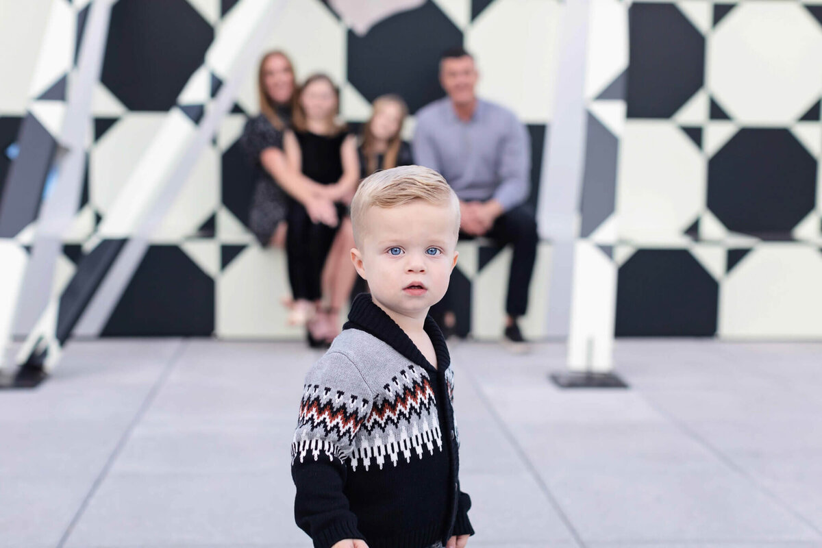 A little boy with blonde hair wearing a grey and black sweater looking at the camera while the rest of his family is sitting far behind him and out of focus up against a wall of black and white circles.
