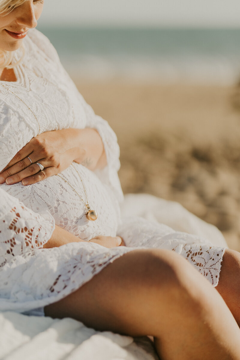 Jeune femme en robe de dentelle blanche, assise regardant et tenant dans ses mains  son ventre rond à la plage. Séance photo grossesse réalisée en Vendée.