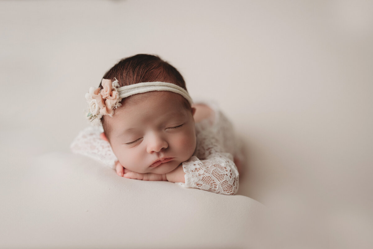 newborn baby girl in cream romper and cream floral headband lying down on cream backdrop posed with her chin on hands