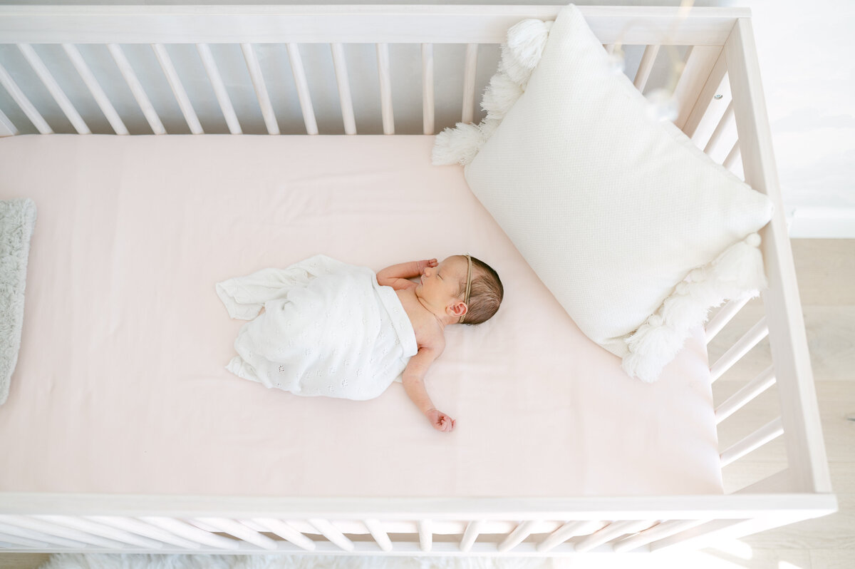 Wide shot from above of a newborn baby laying in her crib and wrapped in a white swaddle with arms out