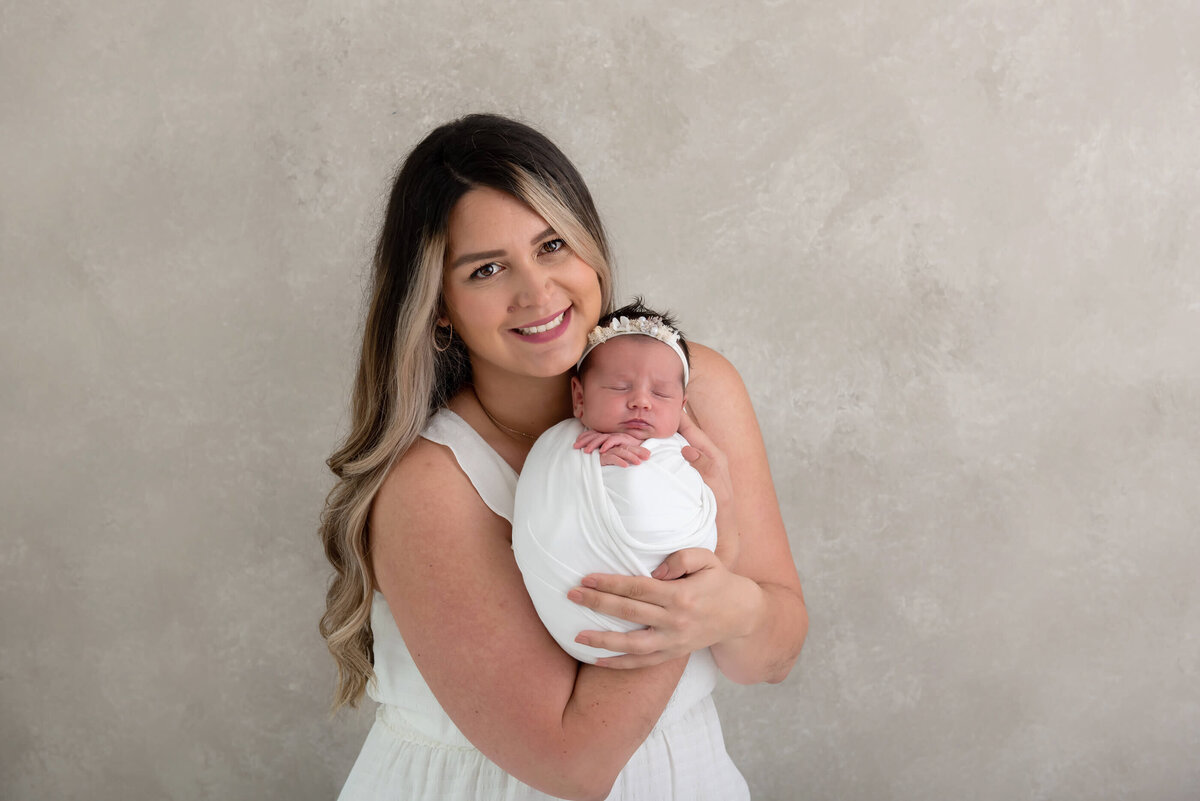 mom holding newborn baby girl wrapped in white for her newborn photos in Burlington, ON