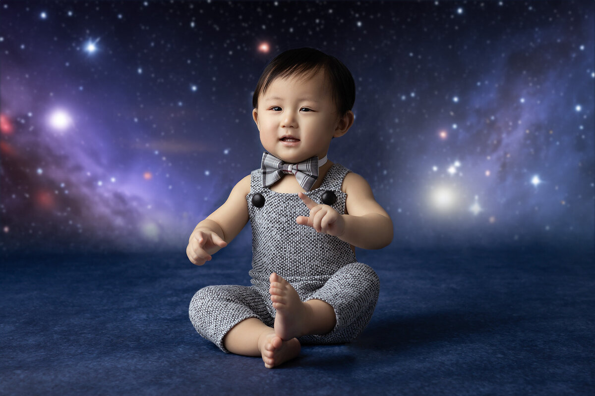 A baby boy in a bowtie and overalls sits with a starry galaxy backdrop, pointing at the sky.