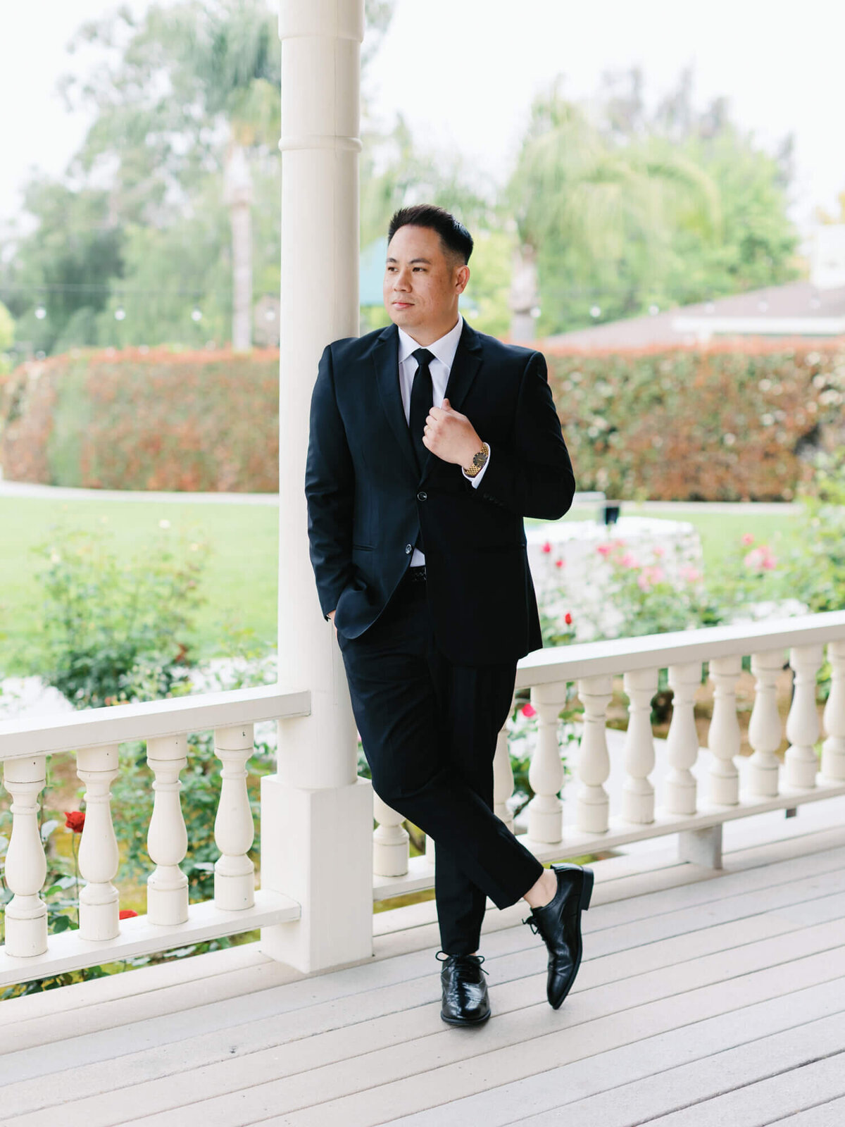 A groom in a black suit and tie stands on a porch, crossing one leg. He gazes into the distance with a thoughtful look.