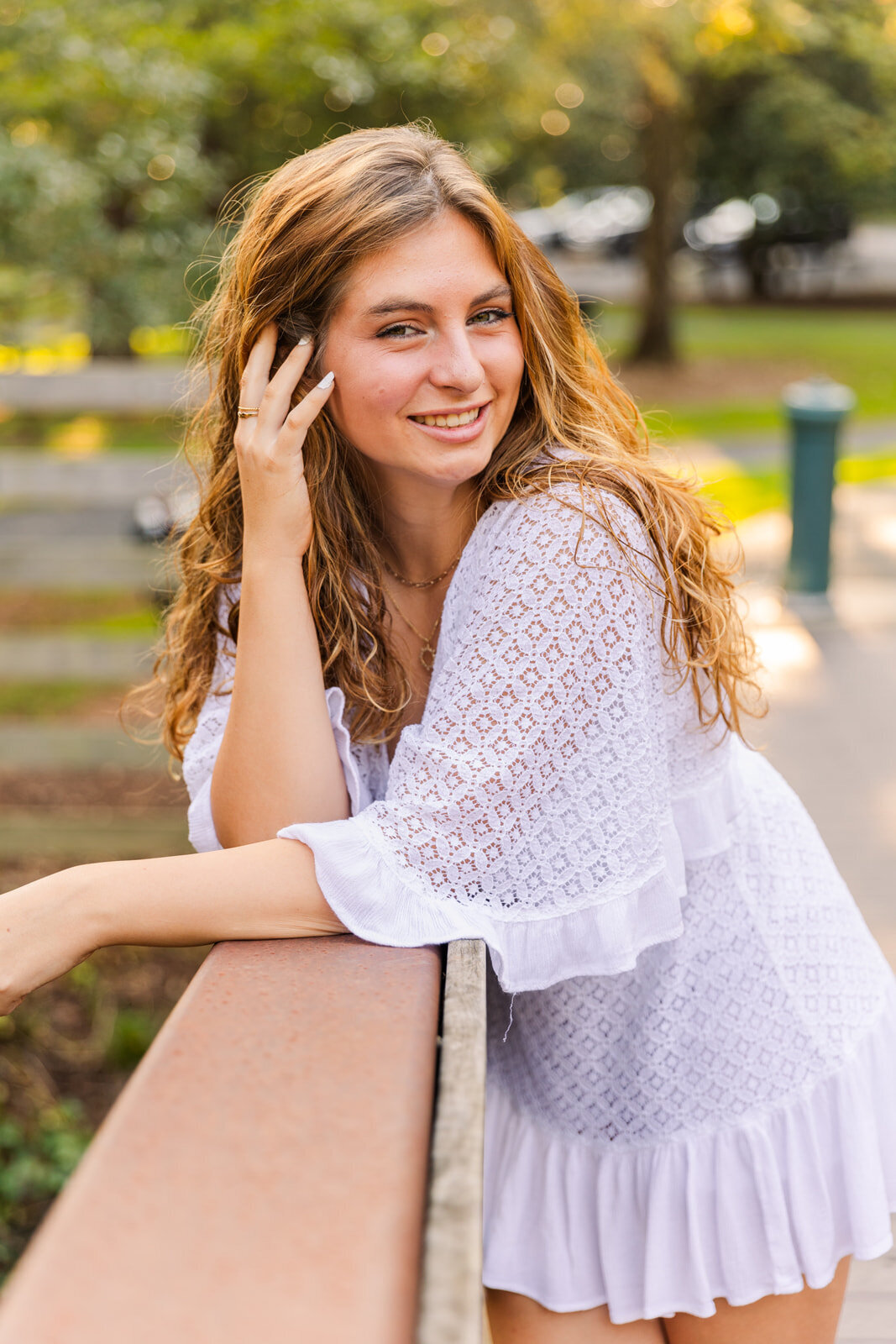 teenage girl standing on a wooden bridge wearing a short white dress resting on the fence her hand in her hair during portrait session