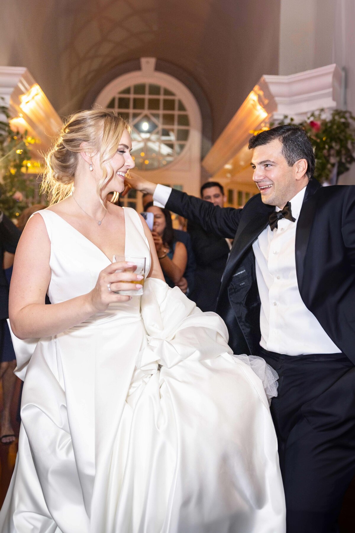 A bride in a white gown holds hands and dances with guests at a formal event. People are wearing formal attire, and theyre surrounded by elegant decor with wooden floors and large windows.