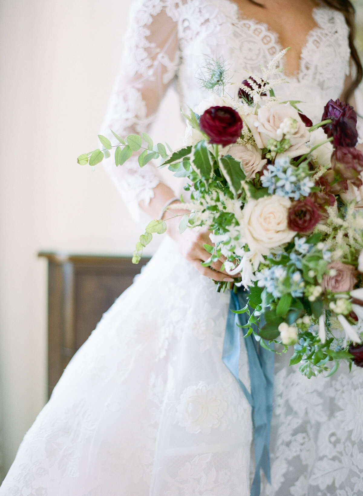 This stunning photo was taken at the Washington National Cathedral in Washington D.C. by photographer Twah Dougherty. In the foreground, a beautiful bride in a white lace wedding dress gracefully holds a large, vibrant bouquet of mixed flowers. The bouquet is a gorgeous combination of roses, greenery, and blue accents, featuring deep burgundy, pale pink, and white blooms with a trailing blue ribbon. In addition, the current films as videographers for this event were Videographers. The background is softly blurred, allowing the focus to remain on the bride and her stunning bouquet.