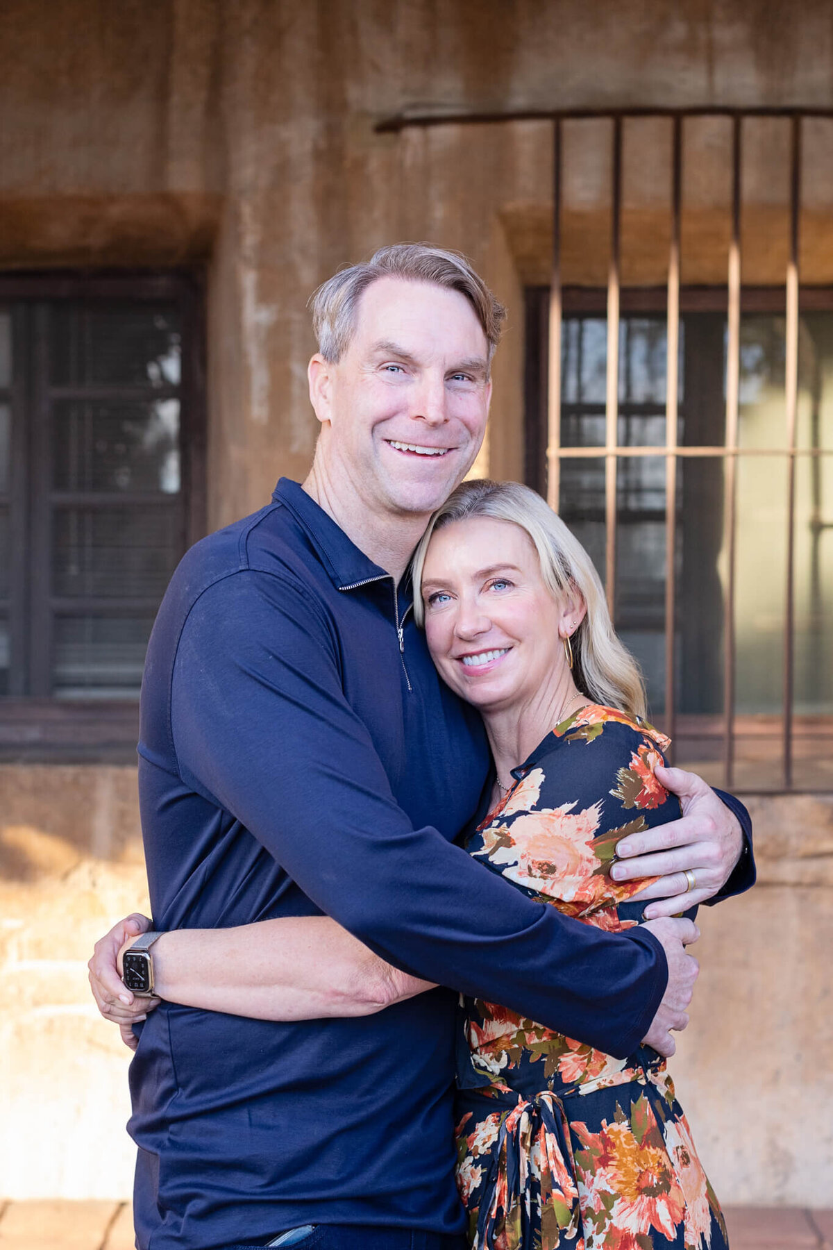 A husband hugging his wife while she is hugging his waist and putting her head on his chest, smiling at the camera.