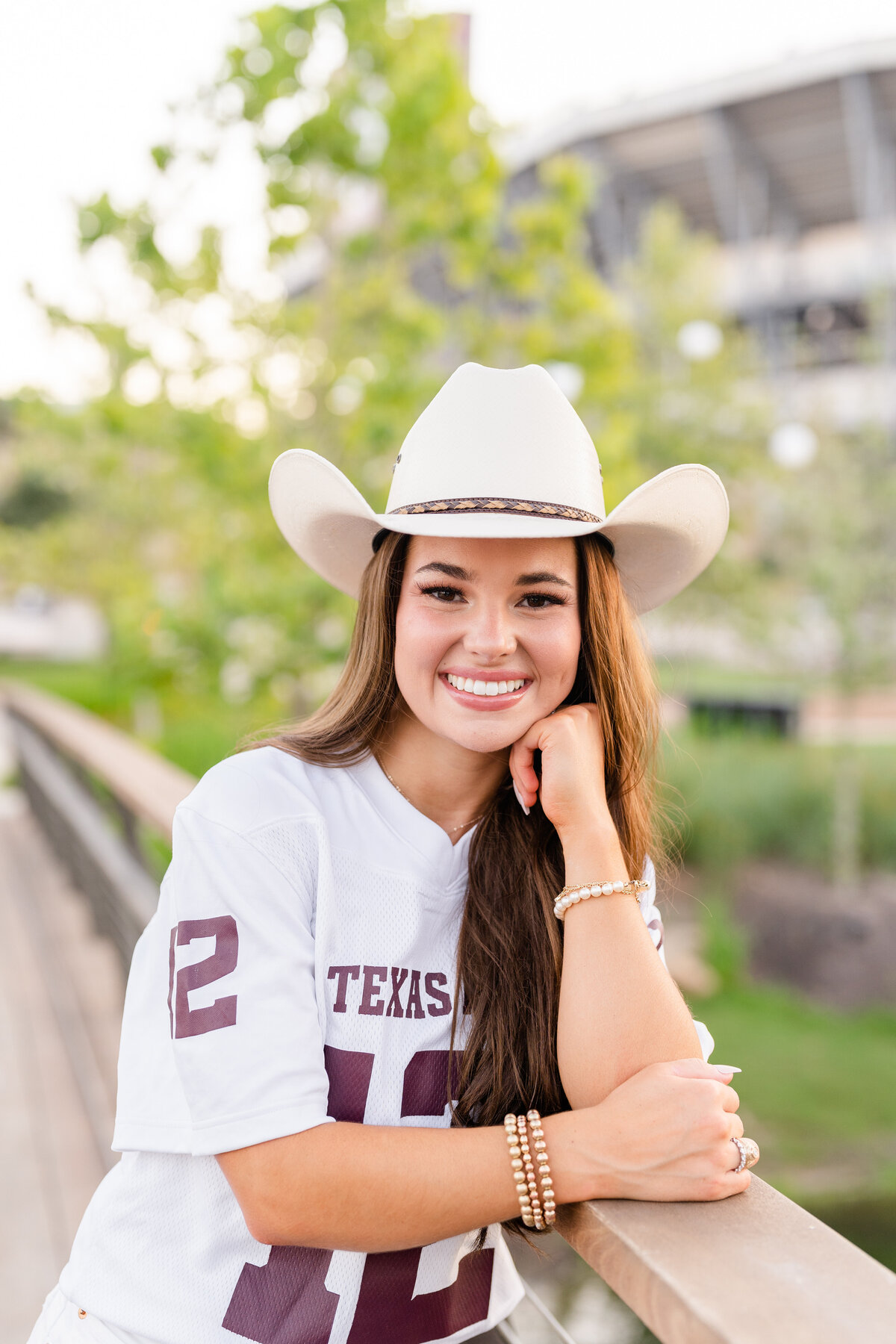Texas A&M senior girl leaning on bridge railing while wearing cowboy hat and white Aggie jersey in Aggie Park