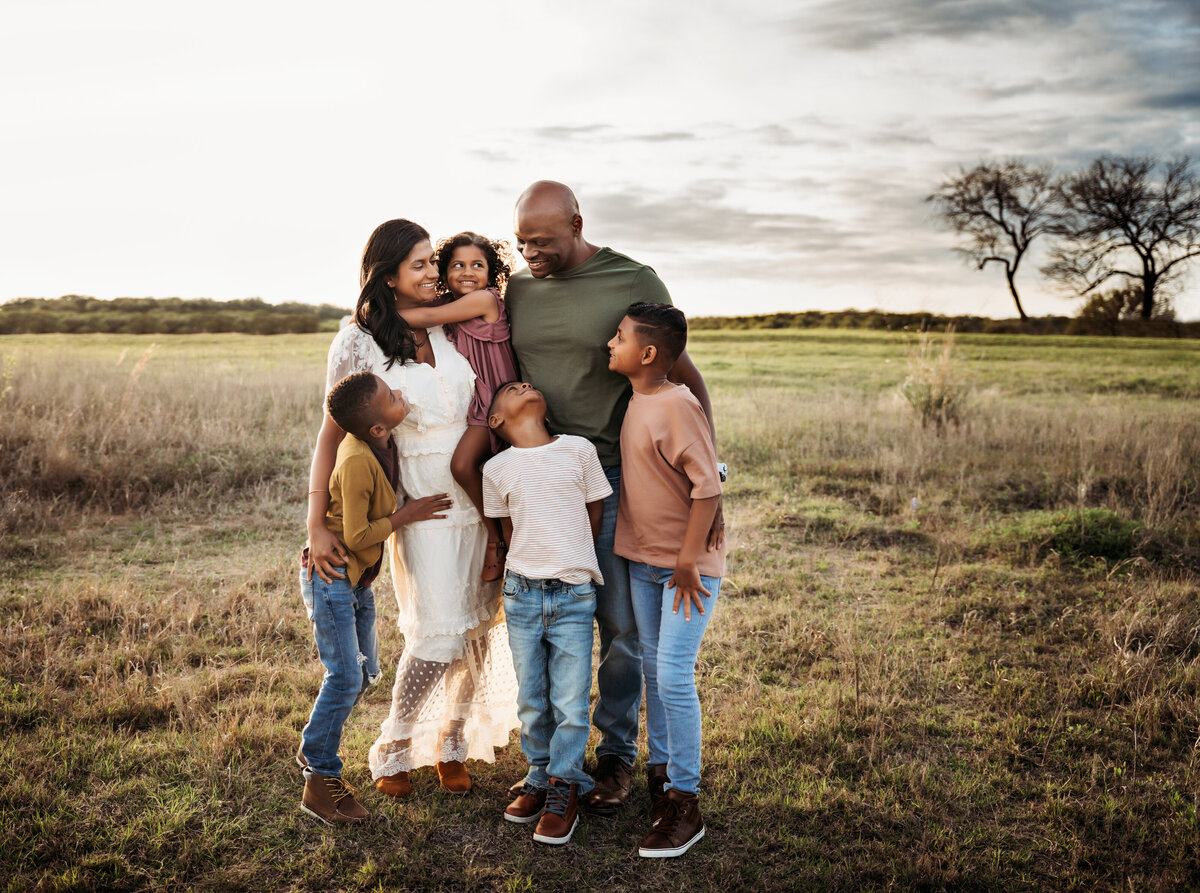 Family Photographer, mom, dad and their four kids huddle close and smiling in an open field