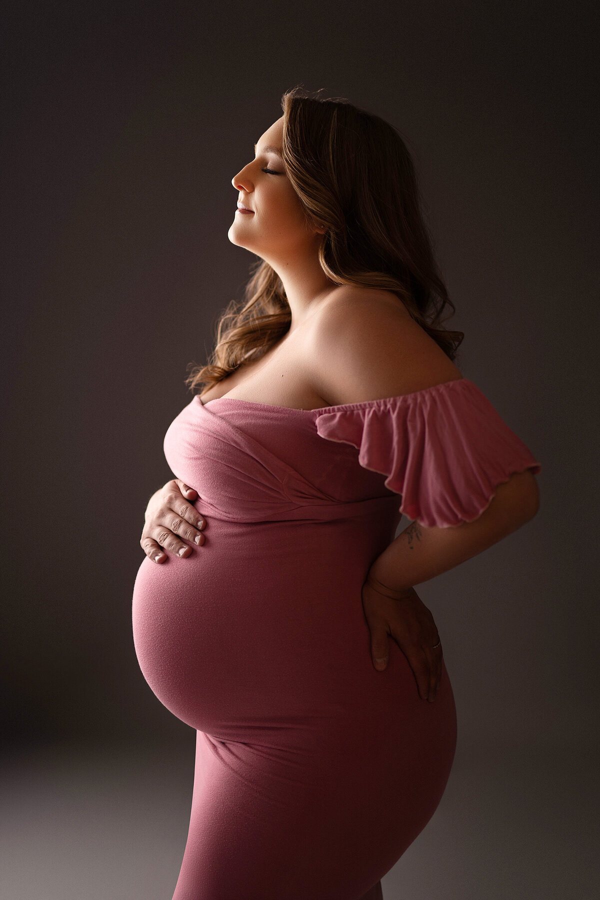 Profile of a pregnant woman wearing a pink, off the shoulder, bodycon maternity gown. Portrain was taken in our Waukesha studio.