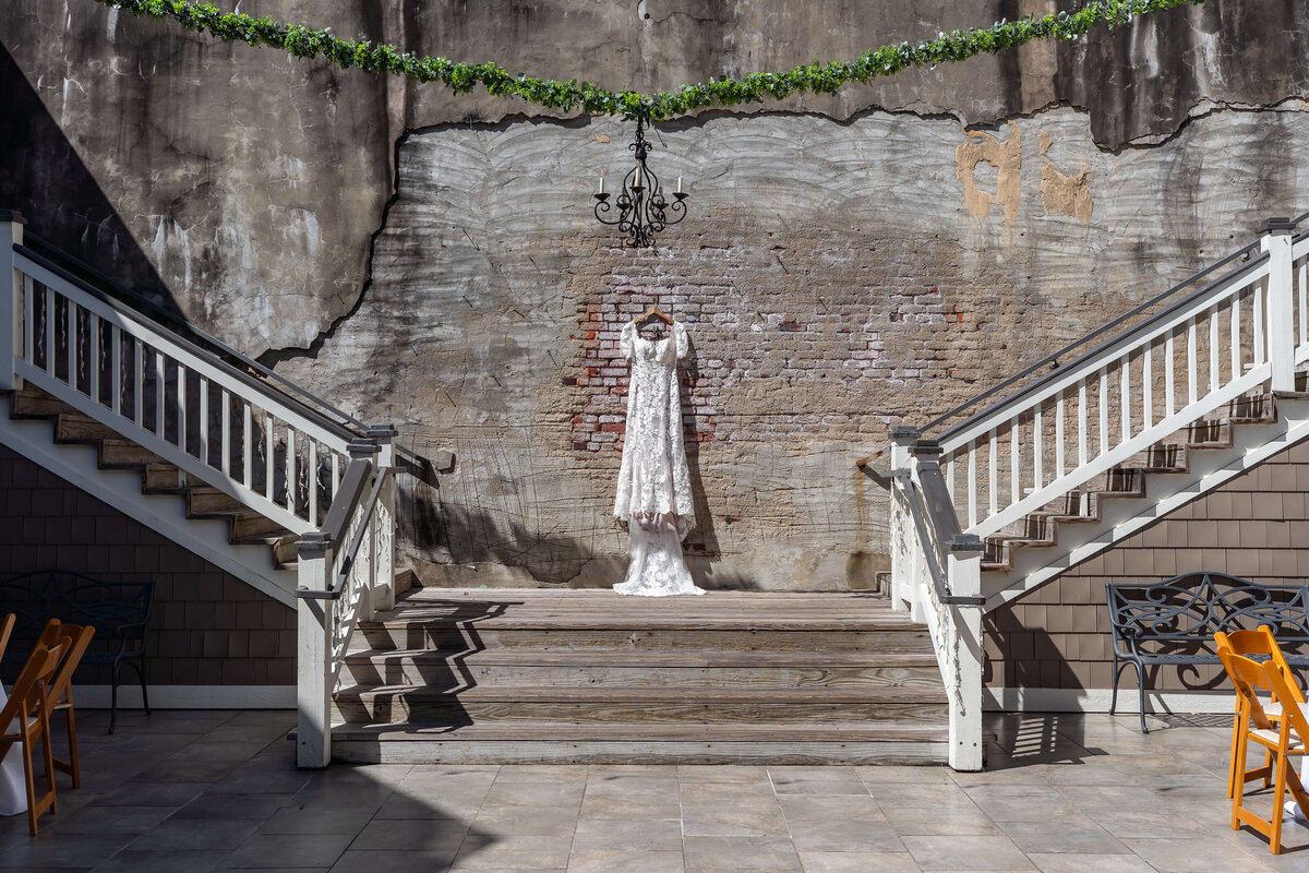 A white lace wedding dress hangs on display between two matching staircases against a weathered brick wall, with a black chandelier and greenery garland above. Sunlight casts clear shadows on the ground.