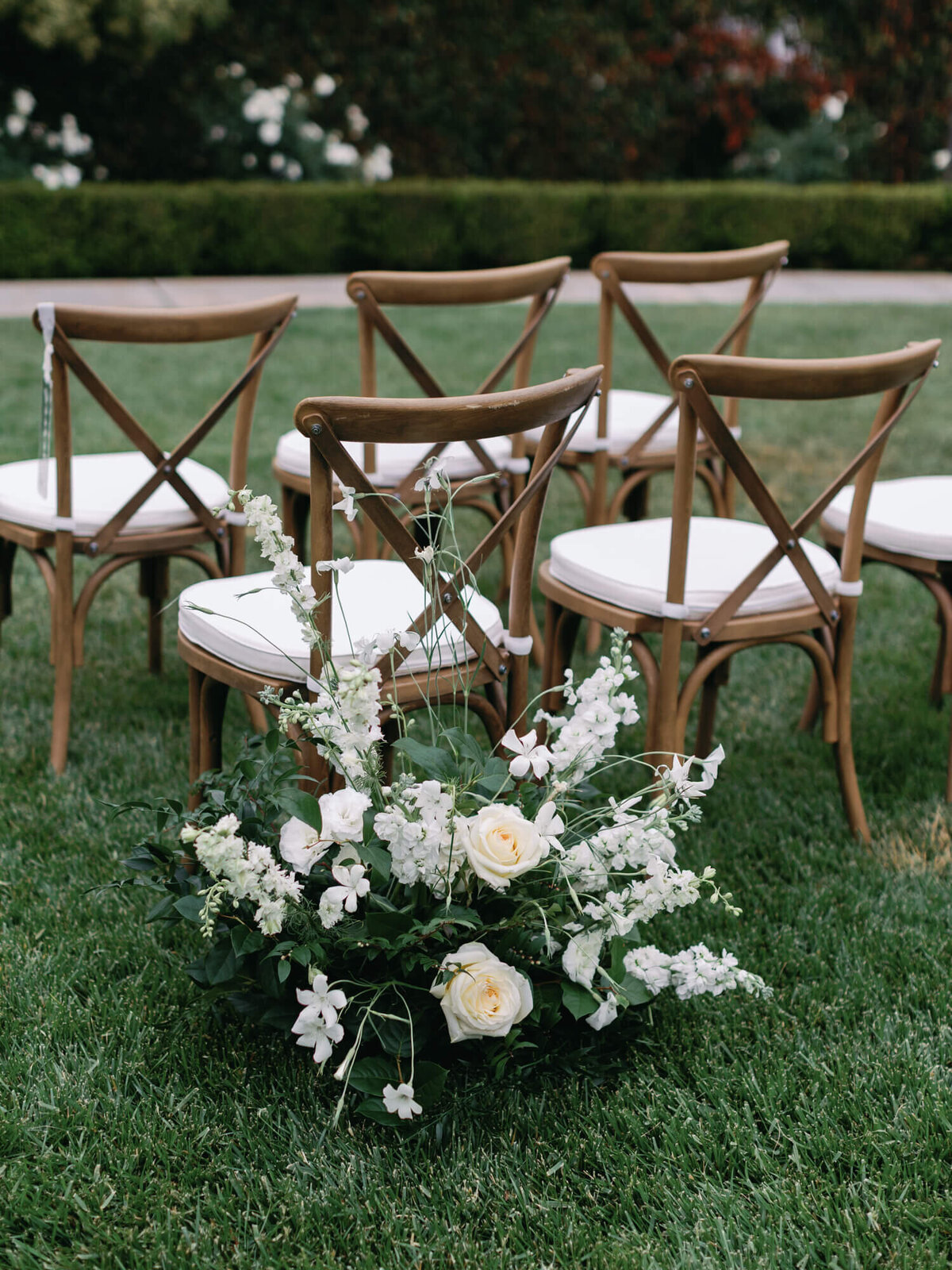 Outdoor wedding setup with wooden chairs and elegant white floral arrangement with roses on green grass in rows.