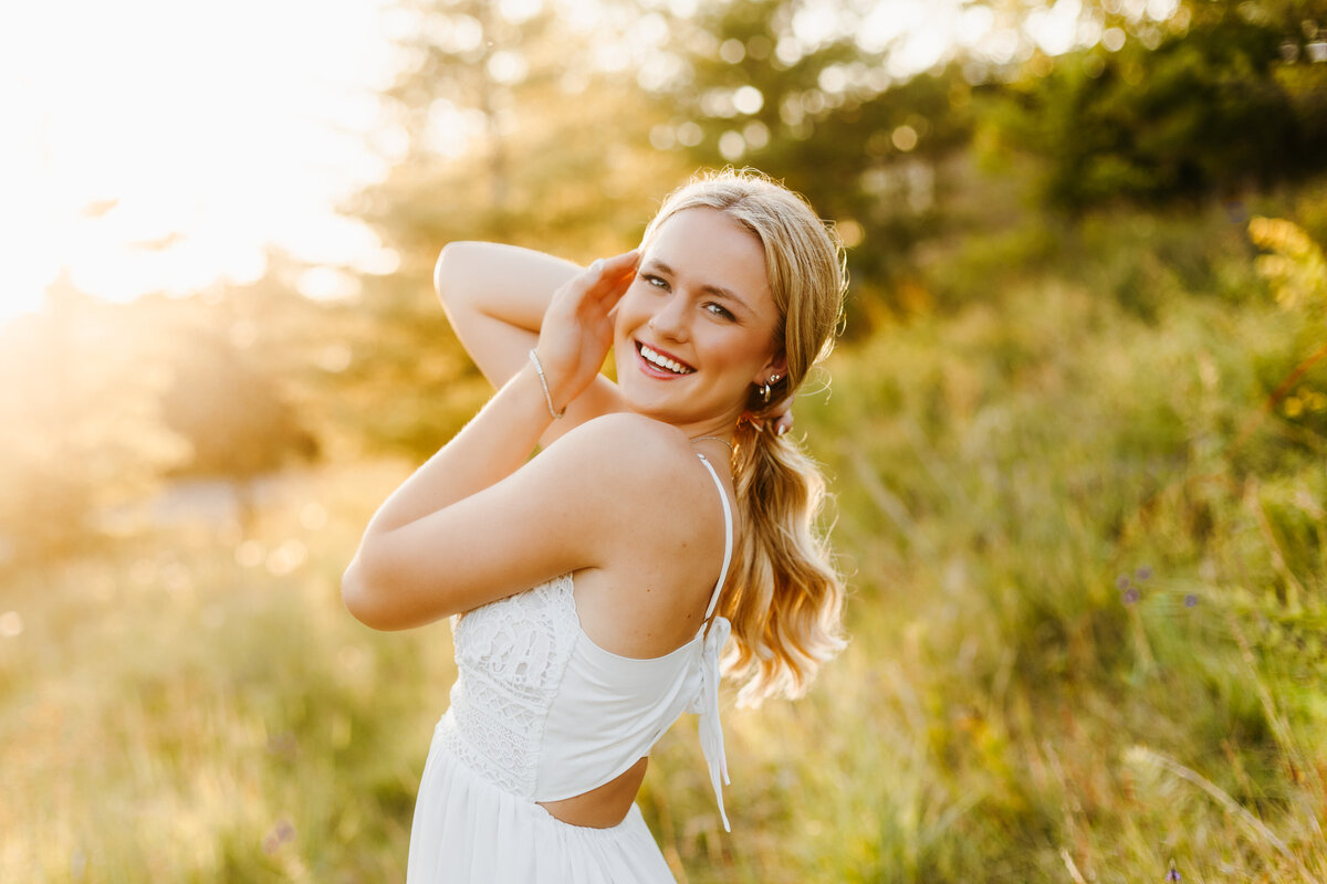 Senior girl in a white dress holding her hair back and standing in a grassy field with the sunset behind her