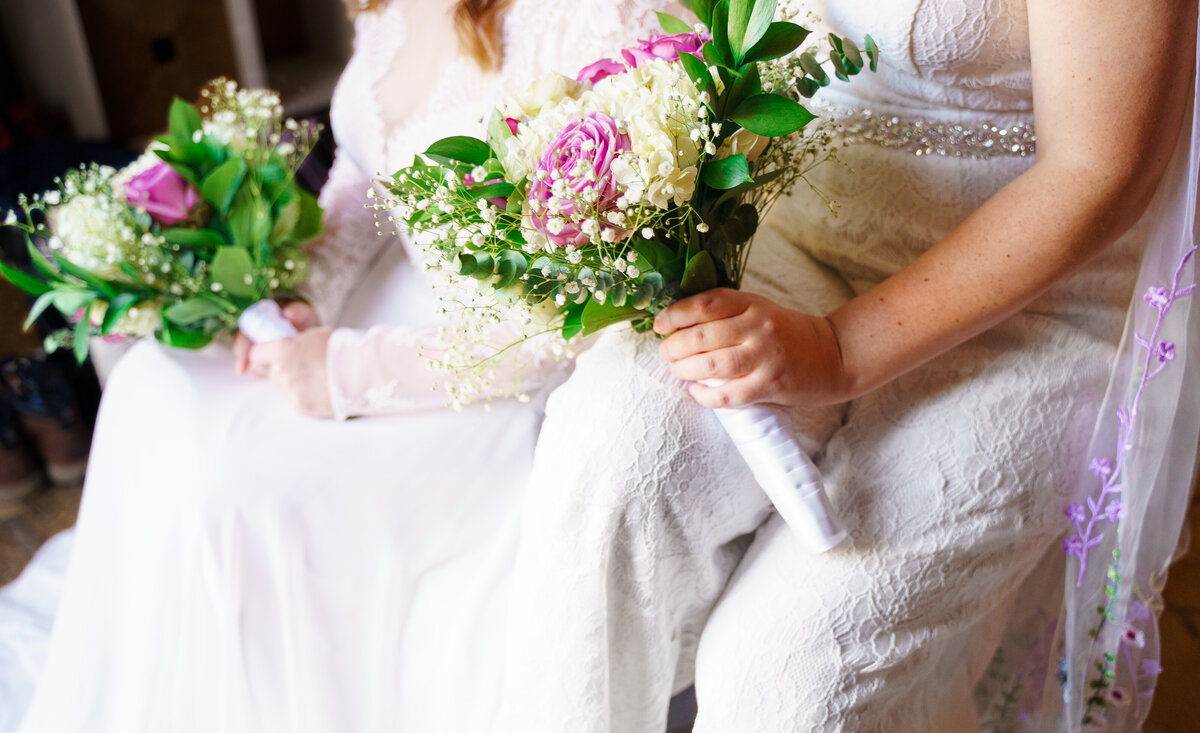 A bride in a white dress and her bride wearing a white jumpsuit hold their bouquets on their laps at their wedding at Cedar Grove Lodging and Events in Hocking Hills.