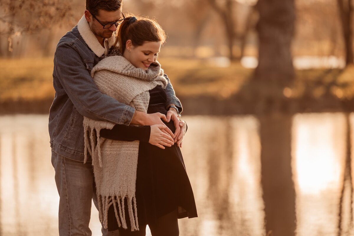 futurs parents en hiver au bord d'un lac et formant un coeur sur le ventre de la femme enceinte
