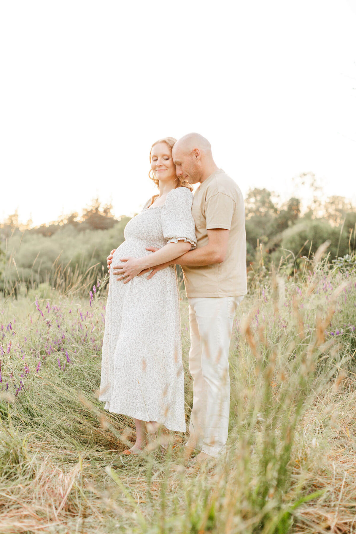 Pregnant woman and man holding her belly and looking down while standing in a beautiful portland natural area at sunset.