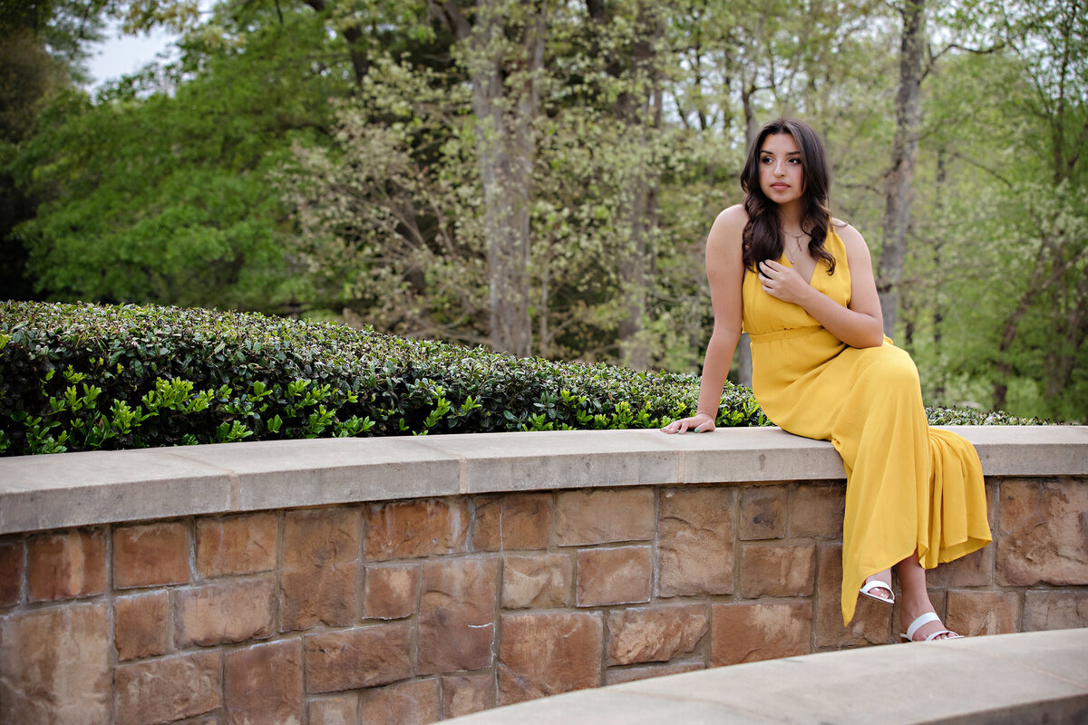 Reflective senior moment in yellow dress sitting on a stone wall with legs dangling, looking off into the distance in a spring garden setting.