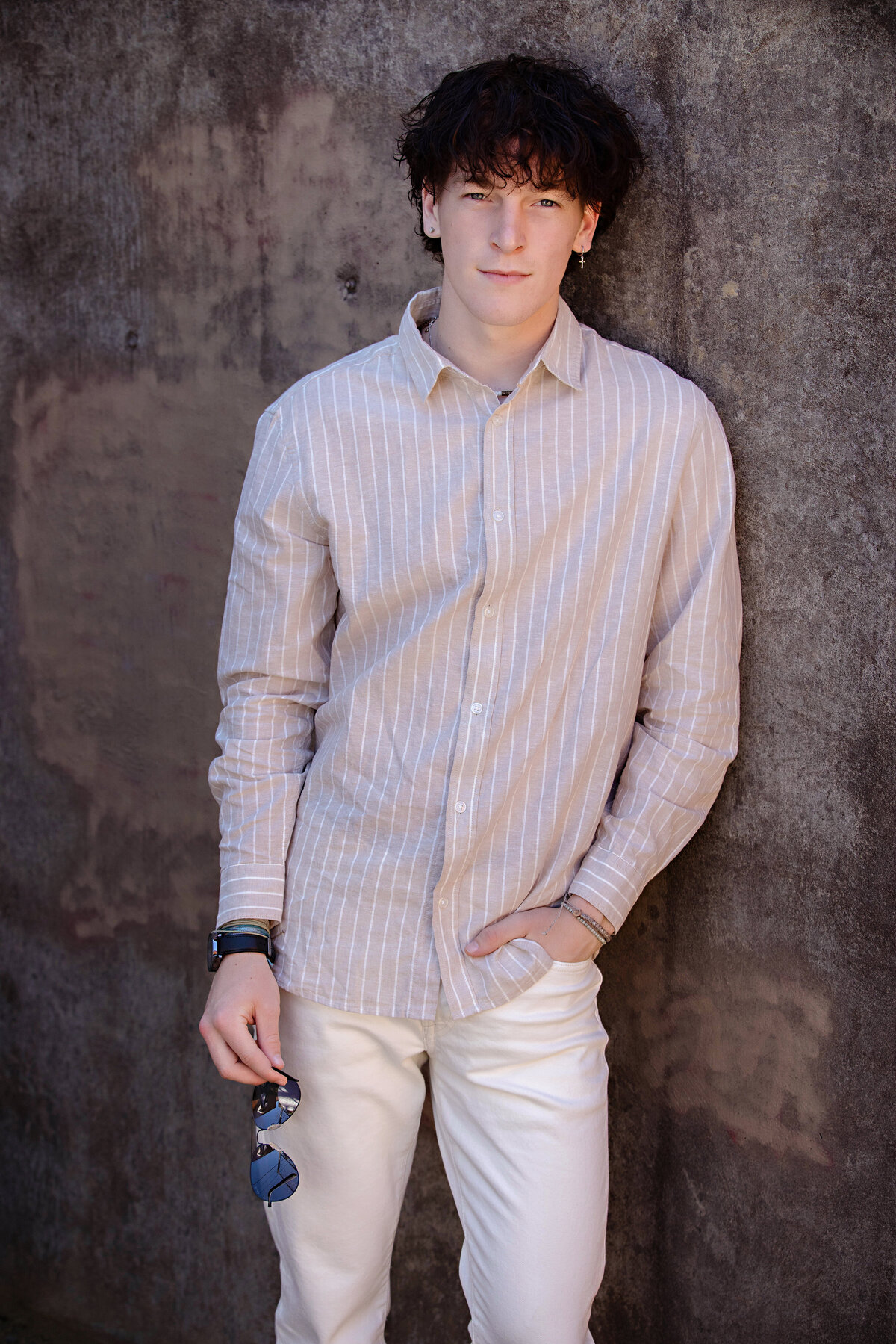 Senior portrait of teen leaning against a stone wall in Charlotte’s urban district, styled in vintage-inspired fashion with sunglasses in hand.