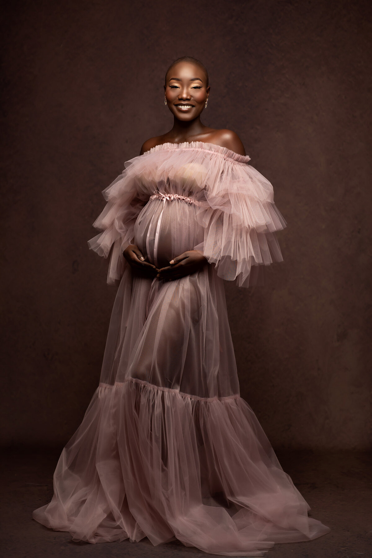 A radiant expecting mother dressed in a delicate, blush pink tulle gown poses gracefully against a warm, textured backdrop. Capturing timeless maternity photography in Dallas, this stunning portrait showcases elegance and beauty.