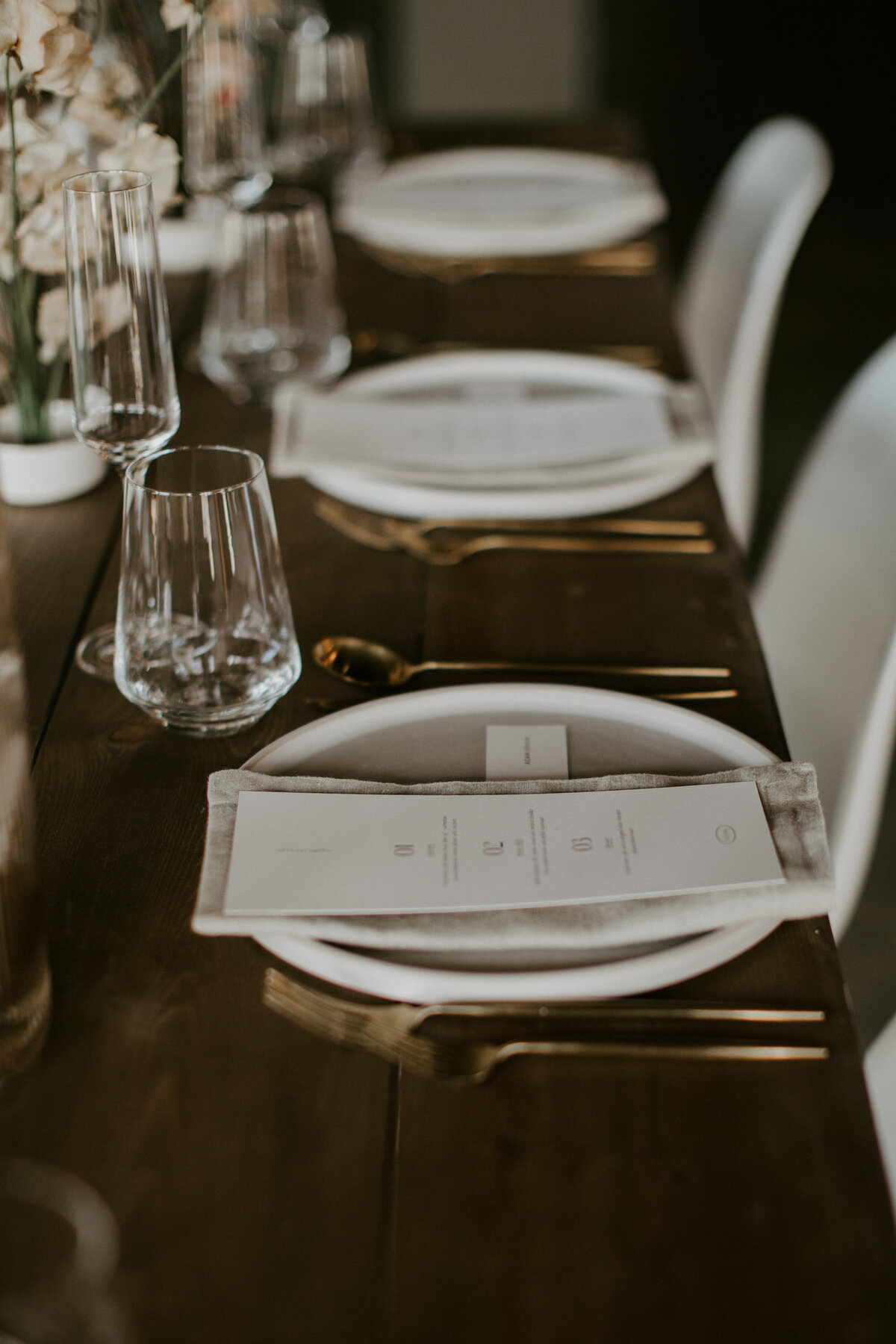 A white menu card with black font atop a linen napkin and white plate set on a wooden table with gold silverware and flowers.