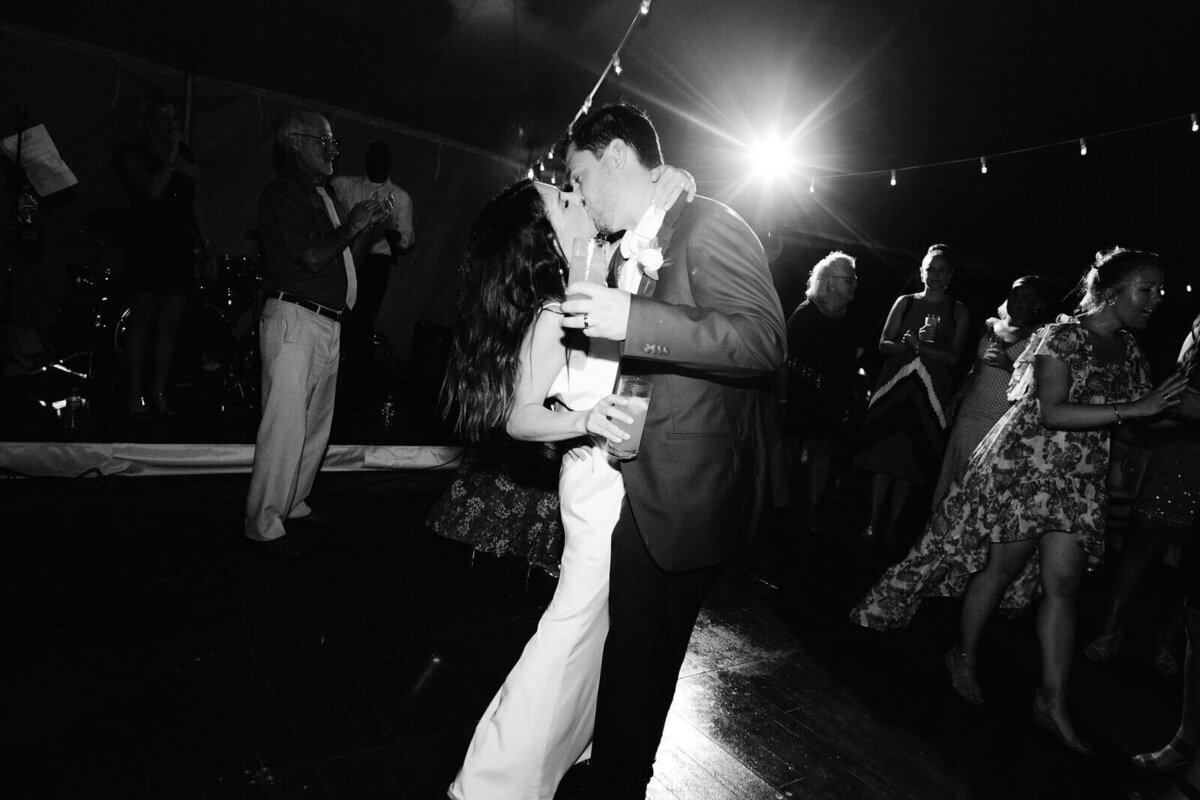 The bride and the groom are kissing each other on the dance floor, as the guests watch on, in Cape Cod Summer Tent, MA.