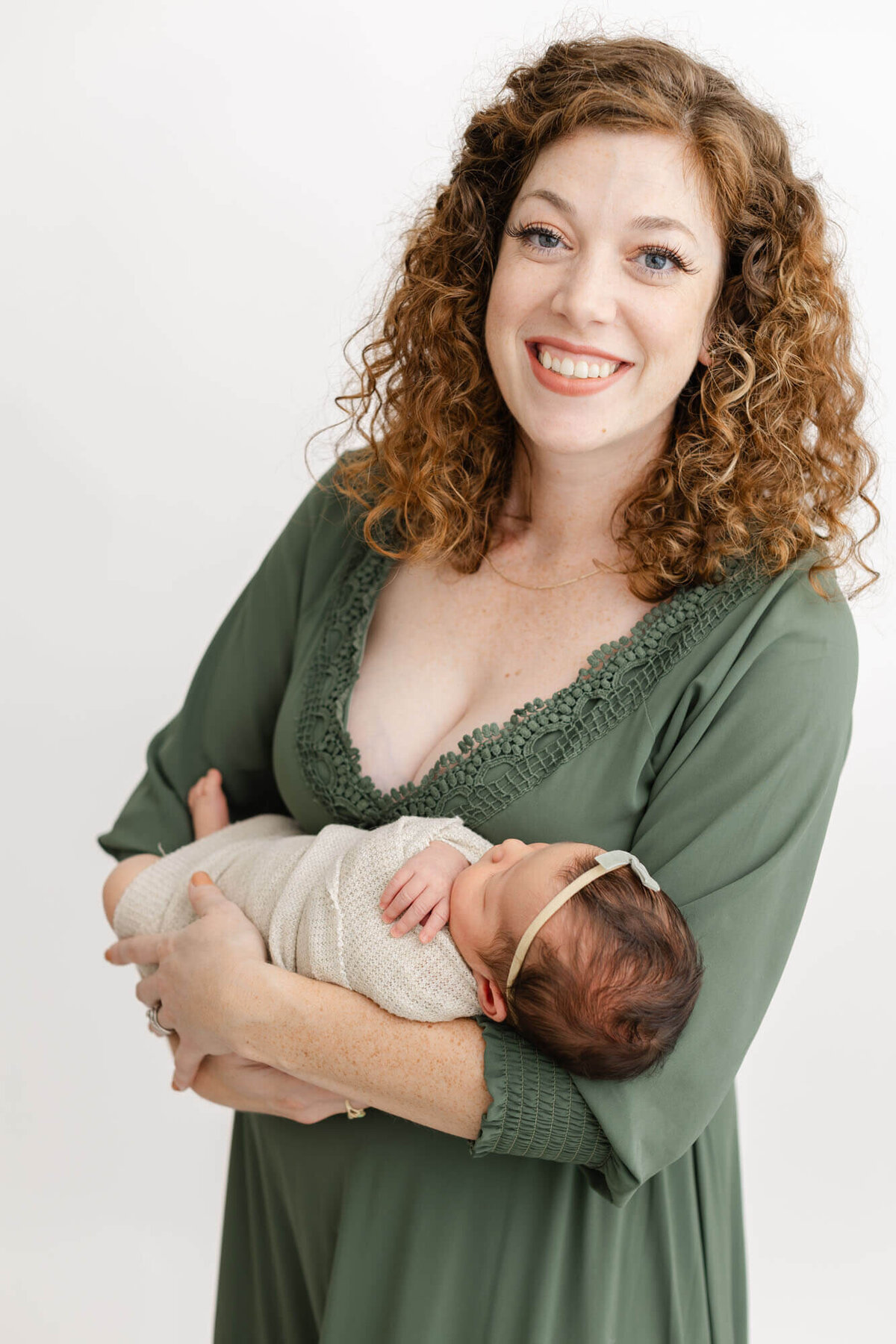 Mom in green dress is holding baby in her arms and smiling towards the camera.