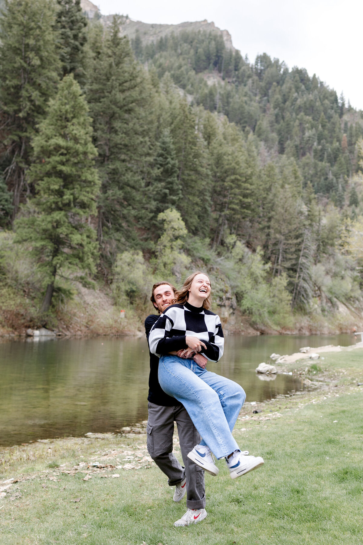 A man playfully lifting his fiancée as they laugh together near a tranquil river at Vivian Park, surrounded by lush forested hills.