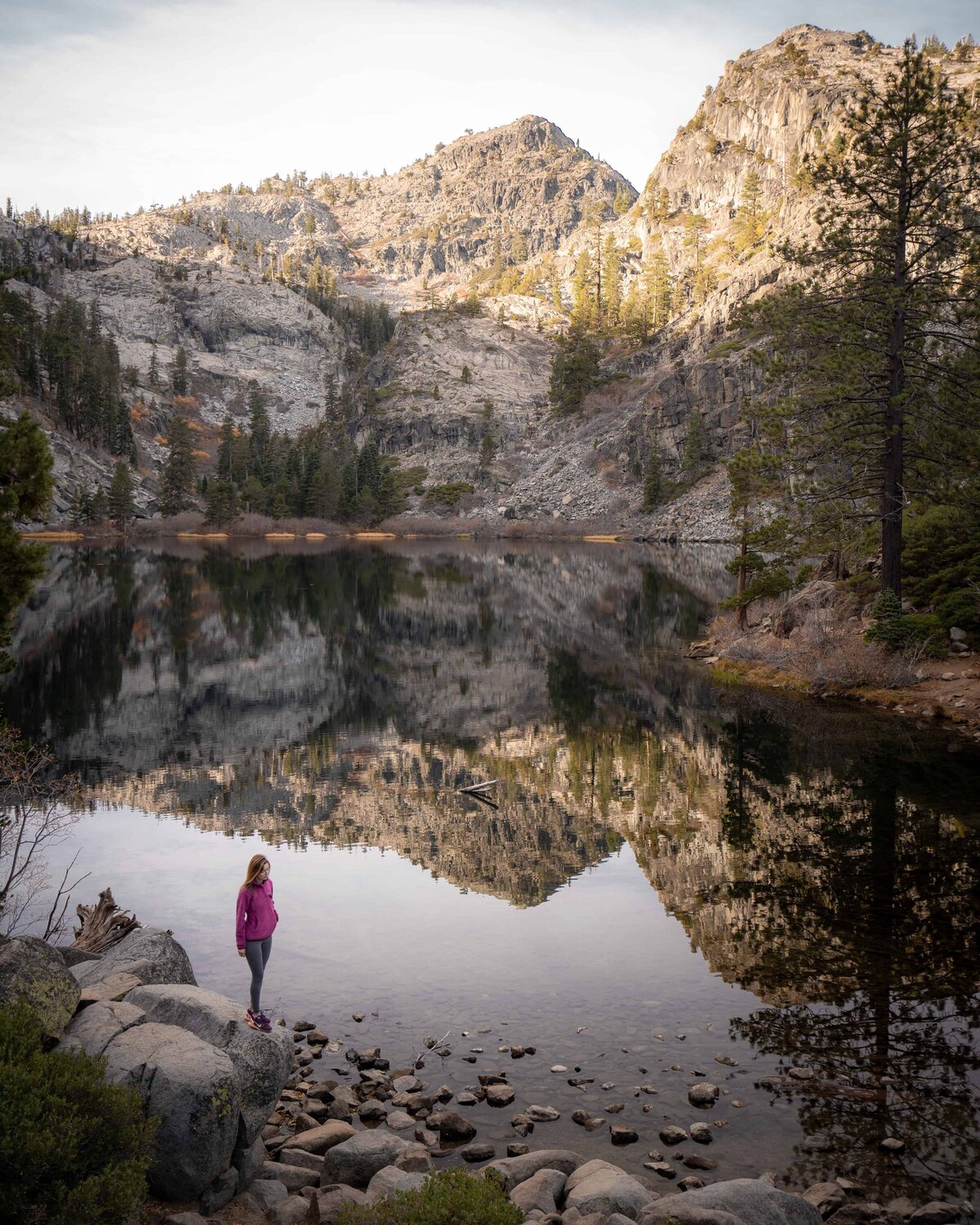 Woman in pink coat standing on rock next to Lake Tahoe with mountains in the background