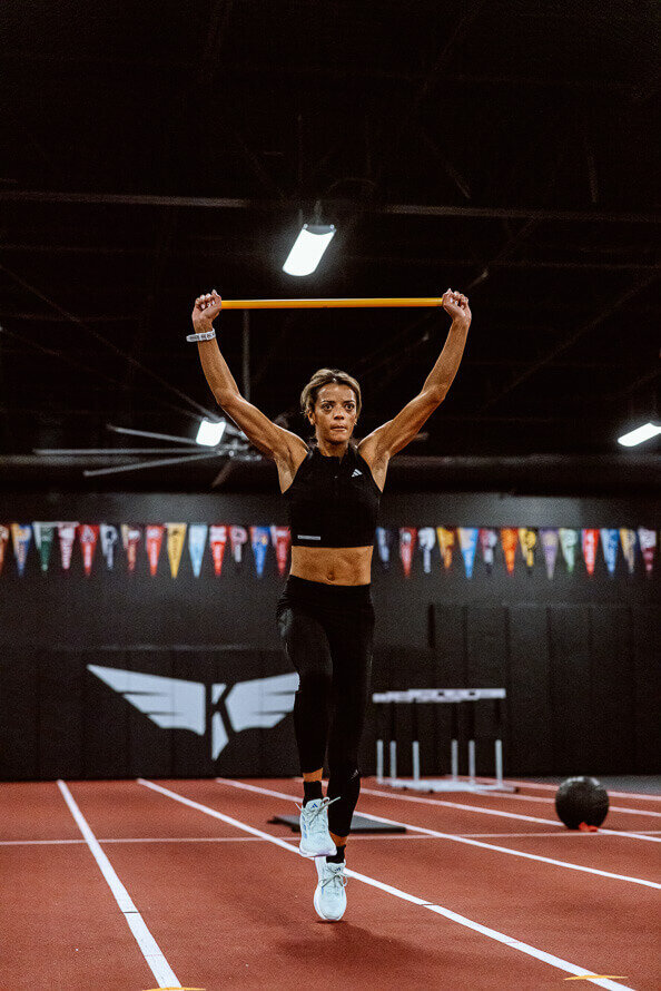 Pro-runner Kaela Edwards holds bar above head while training at the gym.