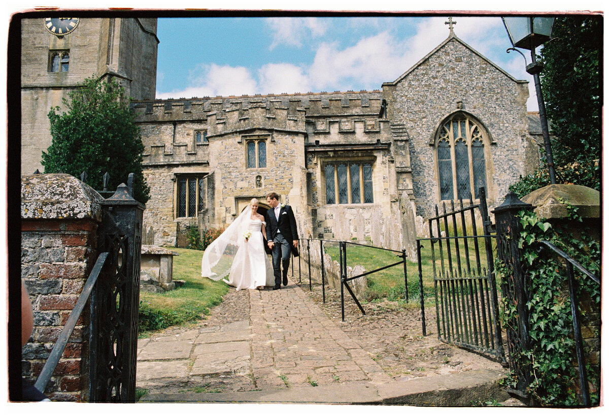 Bride and groom leaving a historic church in rural England, captured on film with fairytale charm and timeless romance — by Kate Winters Photography.