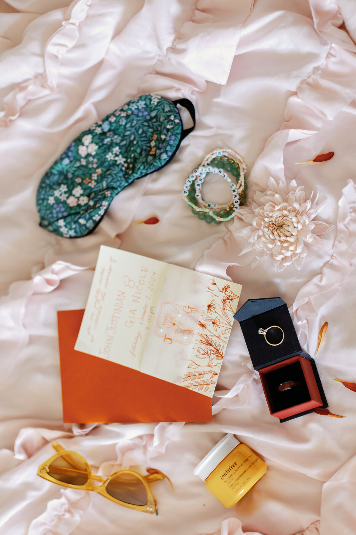 Flatlay of wedding details including invitation suite, jewelry, perfume, sunglasses, and engagement ring on soft pink fabric.