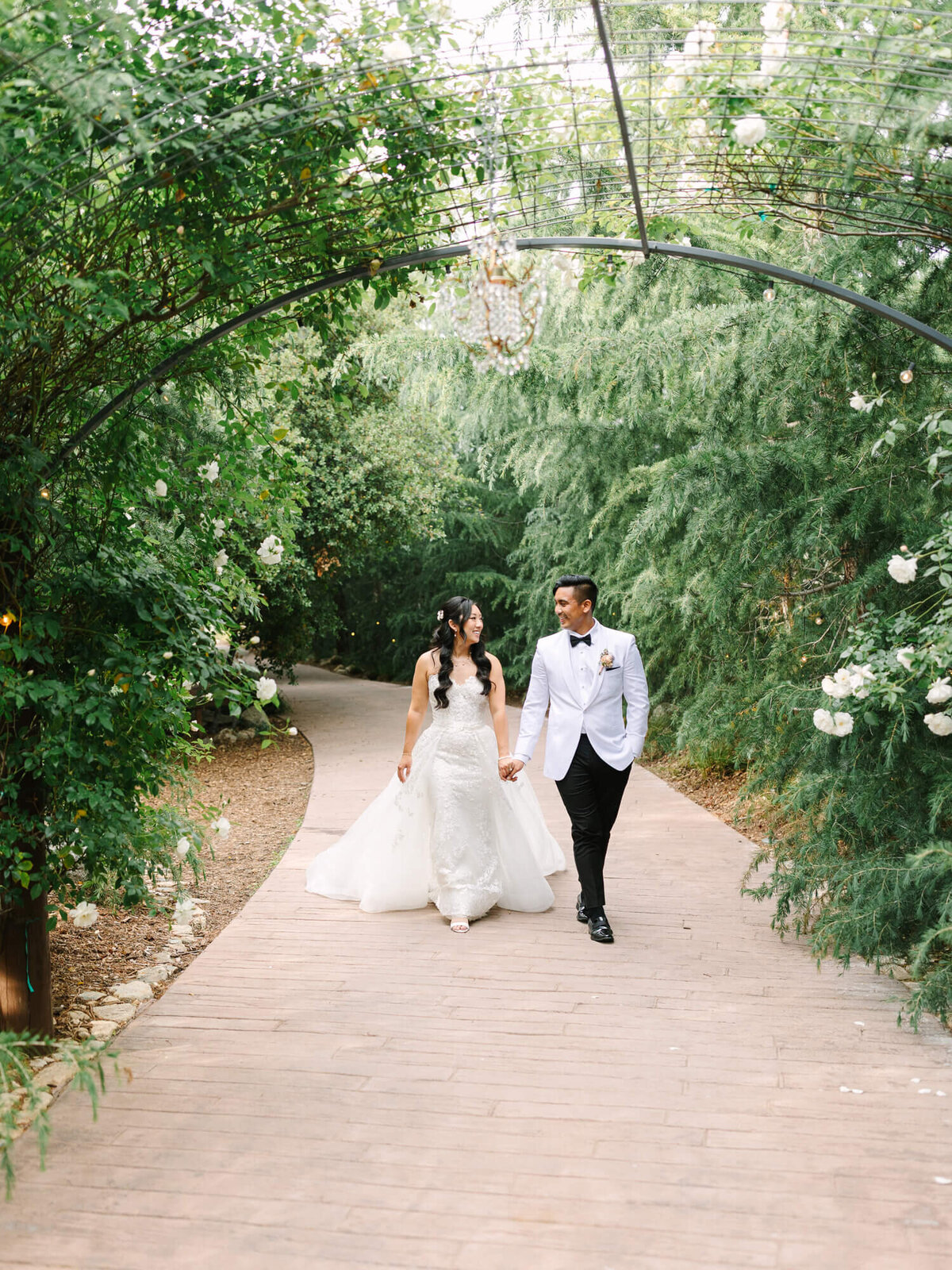 A bride and groom walk hand in hand down a garden path. She wears a lace gown; he wears a white jacket. They're surrounded by lush greenery and flowers.