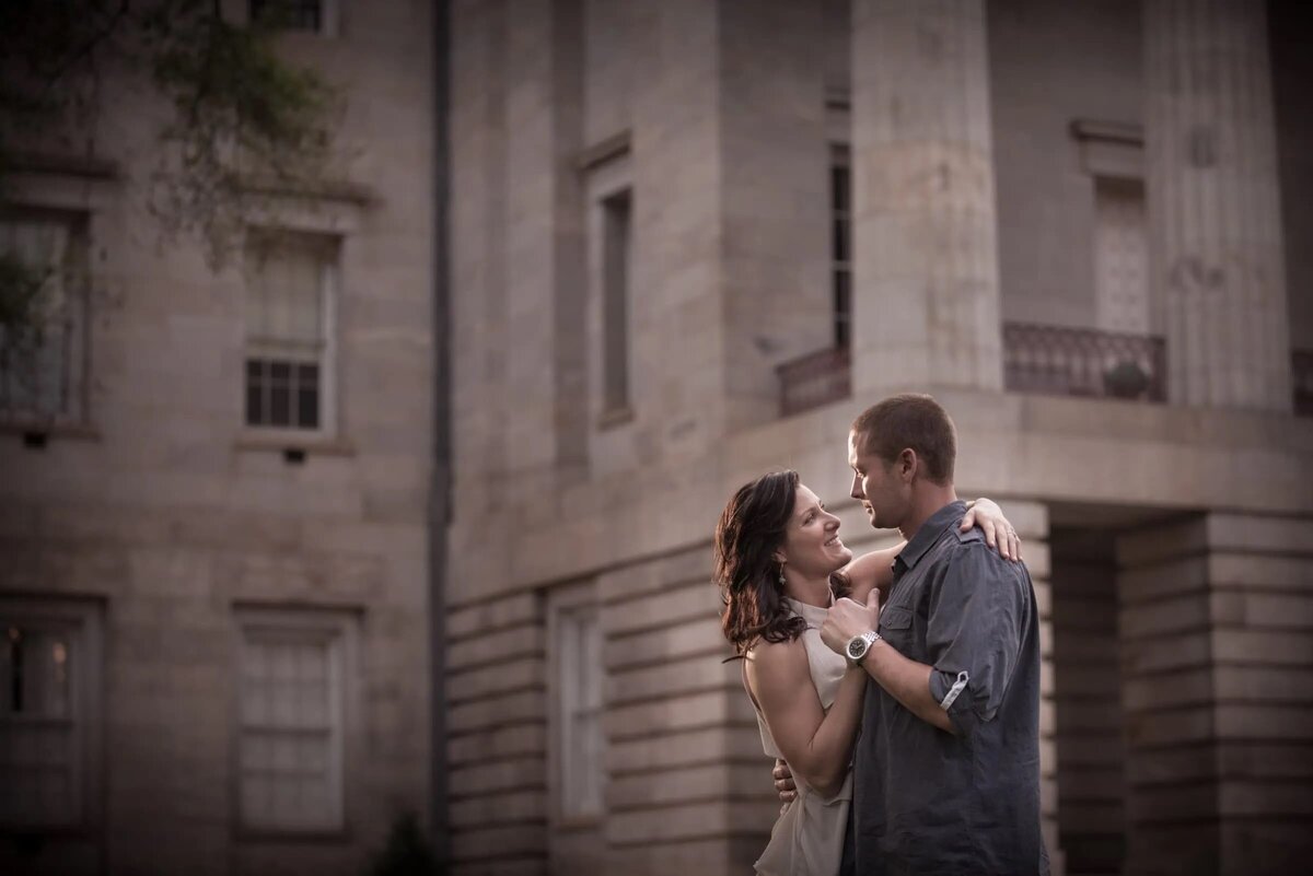 A couple standing in front of a historic stone building, intimately looking into each other’s eyes