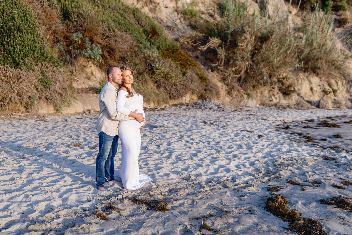 A couple stands on a sandy beach, embraced. The woman wears a white dress, and the man wears a beige jacket with jeans. Behind them are rocky cliffs with greenery. The sun casts long shadows on the sand.