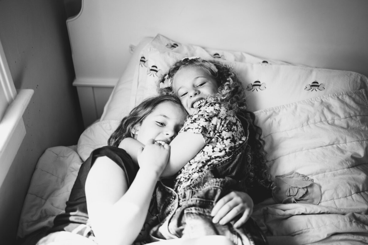 Black & white image of two sisters happily embracing each other in their bed.