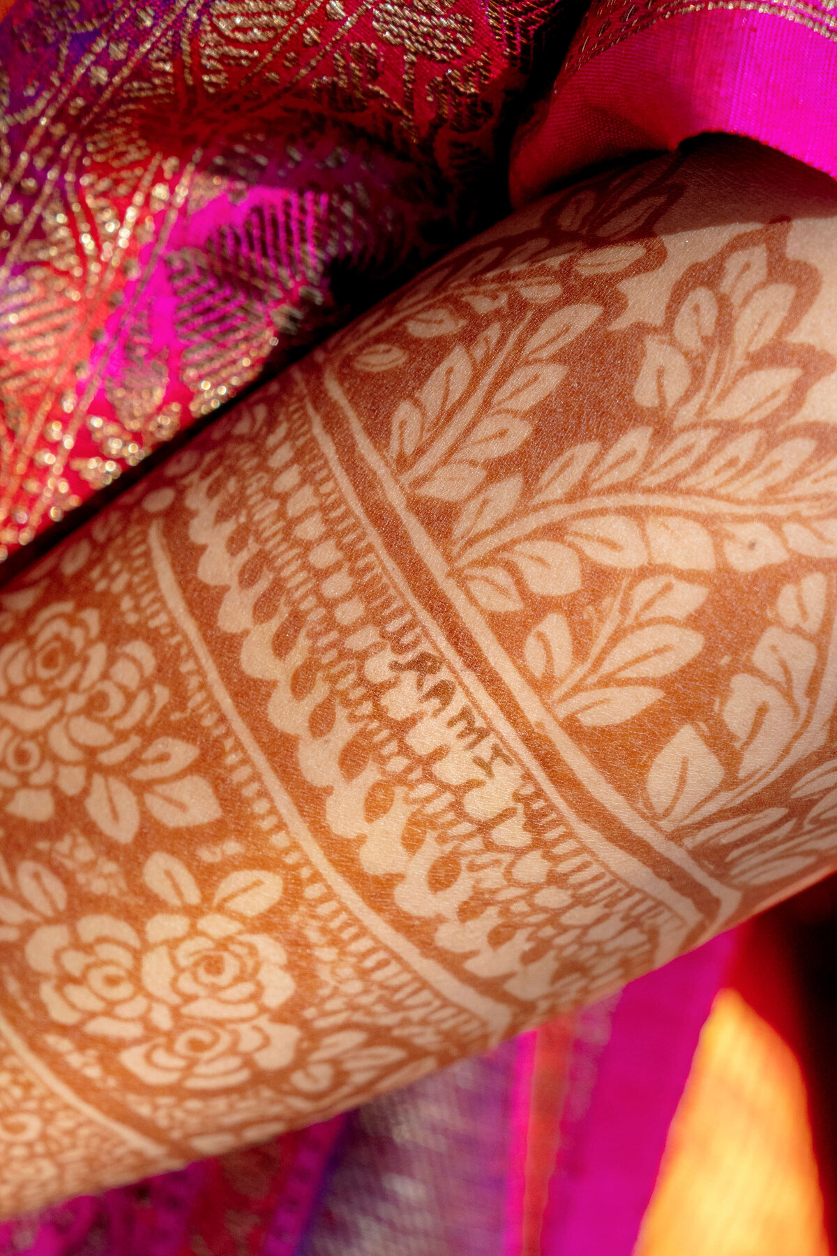Close-up of intricate henna designs featuring floral patterns and leaves on an arm. The background shows a vibrant, shiny pink and gold fabric, partially visible at the top and side of the image.