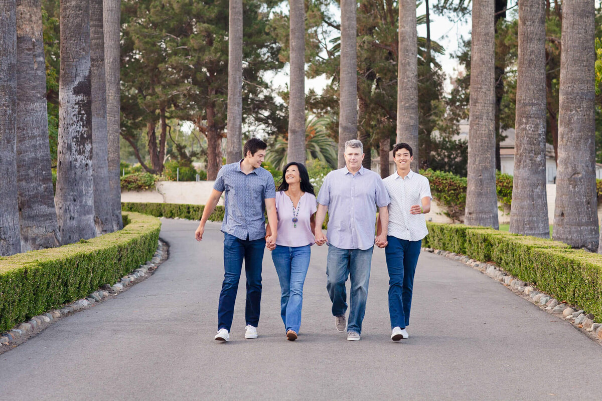 A family of four holding hands and walking towards the camera on an asphalt path with green trees all around them.