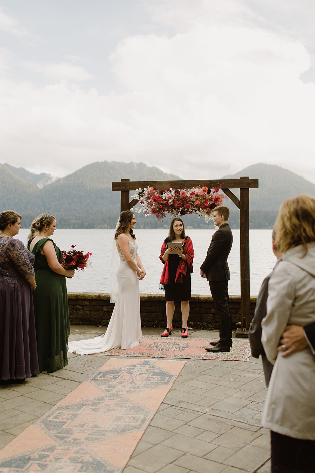 Bride & groom stand with their officiant in front of a wood arch with red, pink & white flowers on Lake Quinault in Olympic National Park!