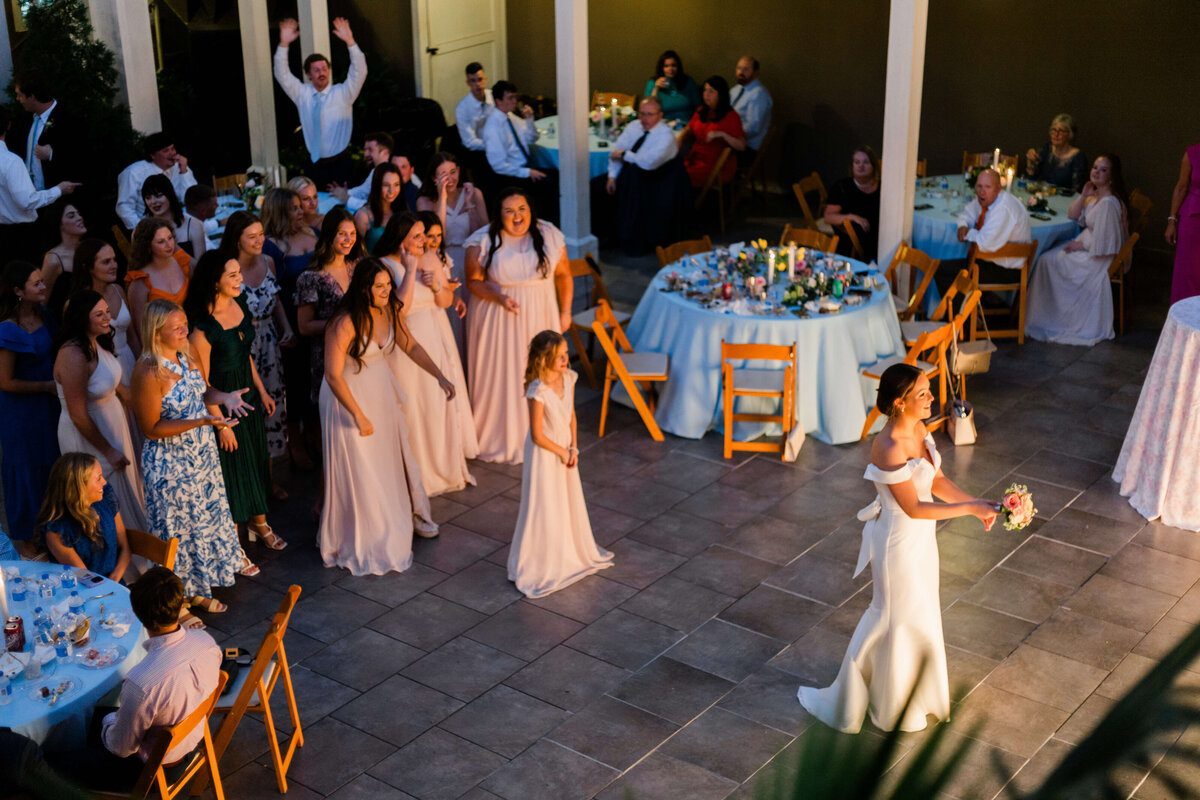 A bride in a white dress prepares to throw her bouquet to a group of excited women and girls gathered behind her at a wedding reception, with guests seated at decorated tables nearby.