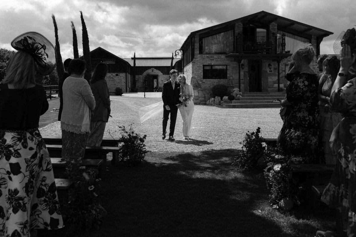 A beautiful black-and-white photo taken at Willow Marsh Farm during the wedding ceremony. The bride and groom, dressed in elegant wedding attire, walk hand-in-hand down the aisle towards their guests. The groom is in a dark suit, while the bride wears a simple, chic white jumpsuit and holds a small bouquet of flowers. The guests, dressed in various formal outfits, look on as the couple walks towards the stunning stone venue in the background. The scene is bathed in soft, natural light, creating a warm and intimate atmosphere.