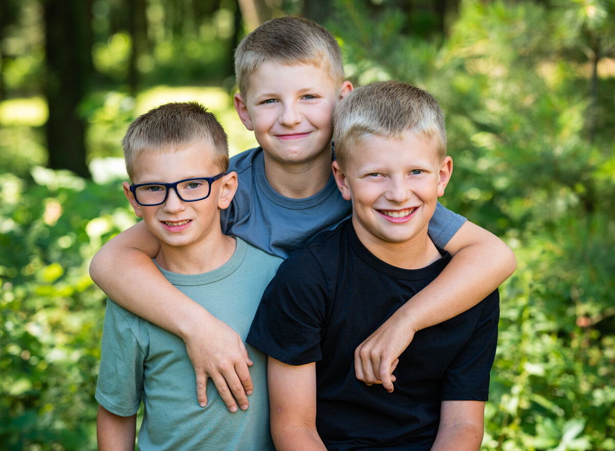 Three young boys smiling and embracing each other outdoors, surrounded by greenery and natural light